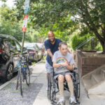 Charles is standing behind Andrew who is using his wheelchair. Charles is pushing the wheelchair uphill on a sidewalk and smiling at the camera. Andrew is also smiling and looking to the side.