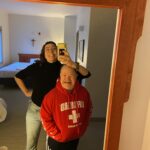 Liddy is standing behind Fritz with her arm resting on his head and her phone camera pointed forward. They're both looking into a mirror smiling in an exaggerated manner. Fritz is wearing a red "Lifeguard" sweatshirt and Liddy is wearing a black quarter length sleeve sweater.
