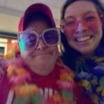 Fritz and Liddy pose for a selfie. Fritz is wearing Elton John type sunglasses, large pink frames with bejeweled edges. He also has a multi-colored flower lei around his neck. Liddy has oversized pink shades on.