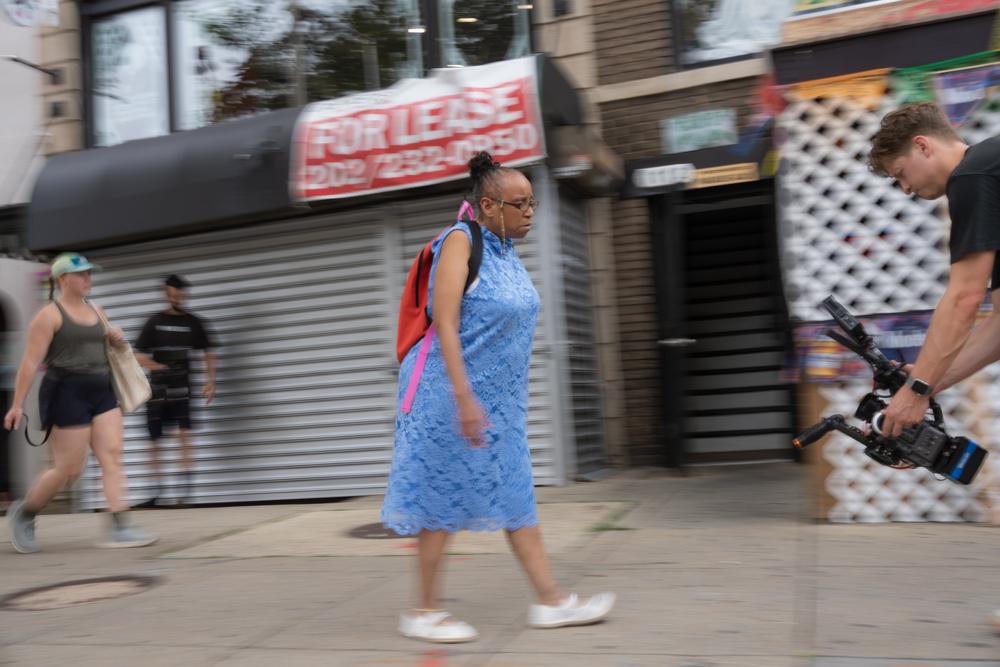 Eileen, a black woman wearing a bright blue dress, white shoes, and a backpack, walks through a city street. Blurs of people are behind her. Zach, a white man with a large camera faces her, walking backwards.