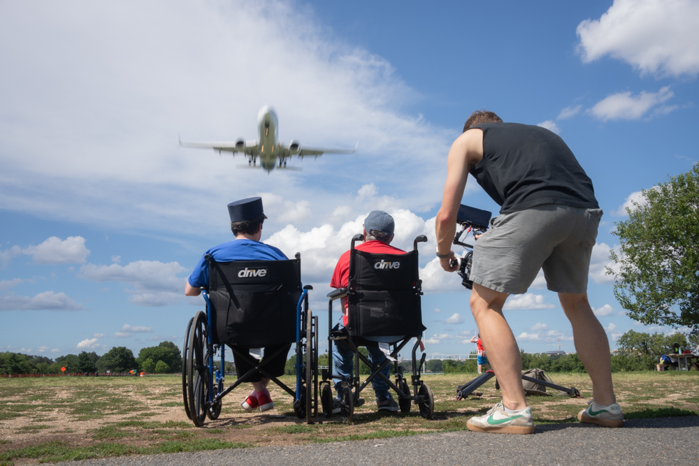 Fran and Bruce sit in wheelchairs at Gravelly Point watching a plane fly low overhead as Zach films them from behind for the Sacred Stories Listening Tour.