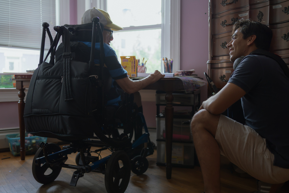 Johnny is sat in his wheelchair in front of his desk, looking down toward Paulo who is kneeling next to him. Both are smiling as they chat.