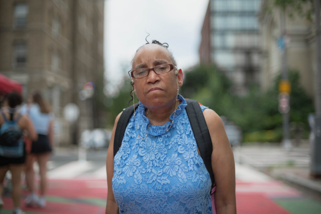 Eileen, a black woman with her hair in high ponytail wearing a bright light blue dress, looks seriously and strongly directly into the camera lens. She's wearing a backpack and posed on a city street.