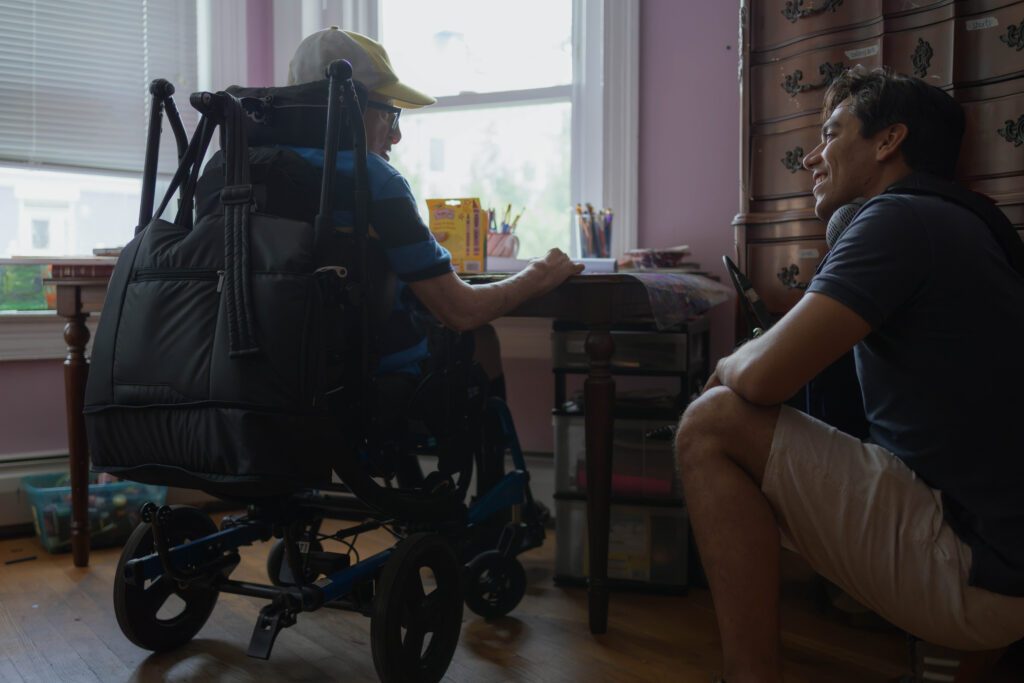 Johnny is sat in his wheelchair in front of his desk, looking down toward Paulo who is kneeling next to him. Both are smiling as they chat.