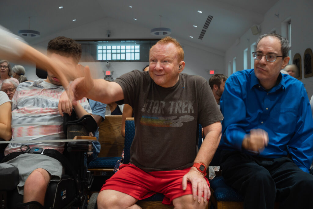 A red-headed, freckled white man named Eric is greeting a passerby who is sort of blurred from their own movement. Eric is sandwiched in a church pew between two other people.
