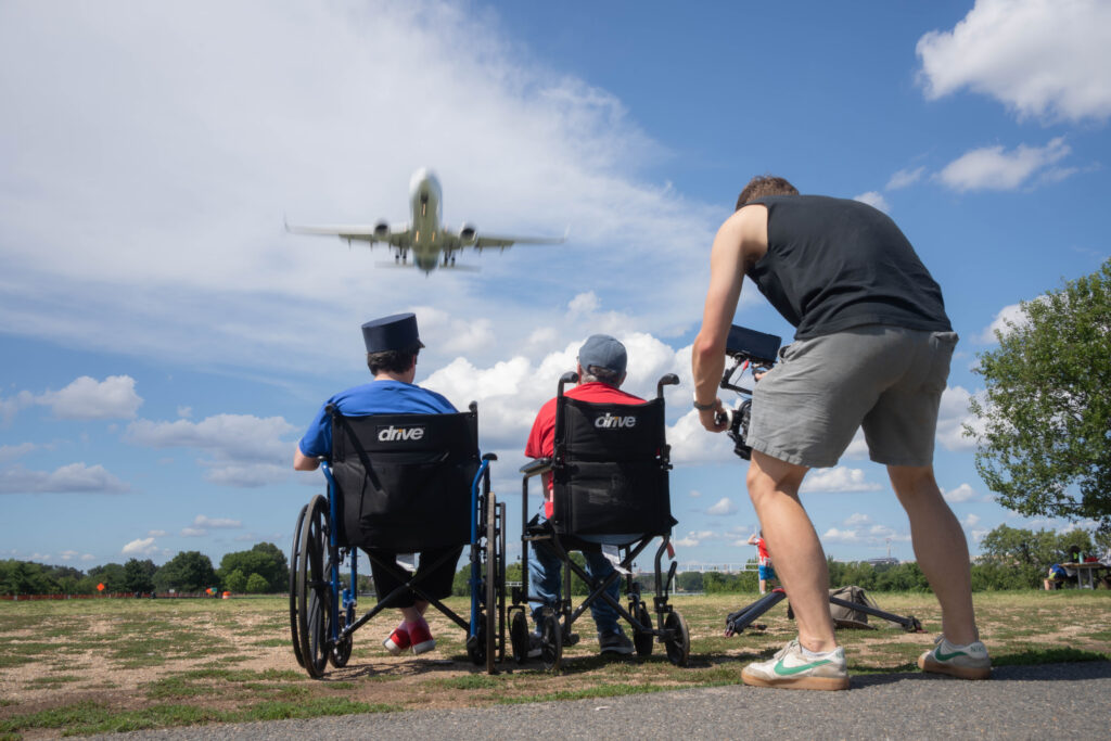 Fran and Bruce are seated in their wheelchairs, backs to us. Zach is behind them with a large camera, filming them watching a huge plane land at Gravelly Point.