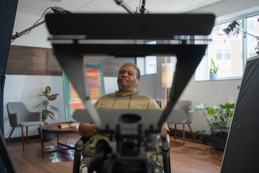 Joseph is framed in a trapezoid-shaped camera, set in an indoor interview room with a colorful painting behind him.