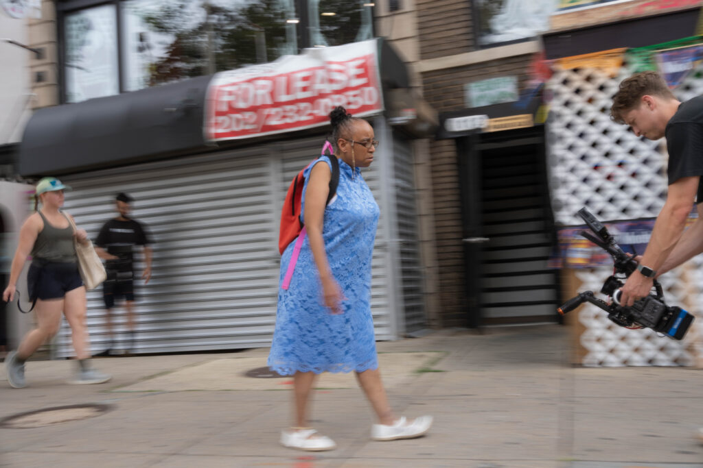 Eileen, a black woman wearing a bright blue dress, white shoes, and a backpack, walks through a city street. Blurs of people are behind her. Zach, a white man with a large camera faces her, walking backwards.