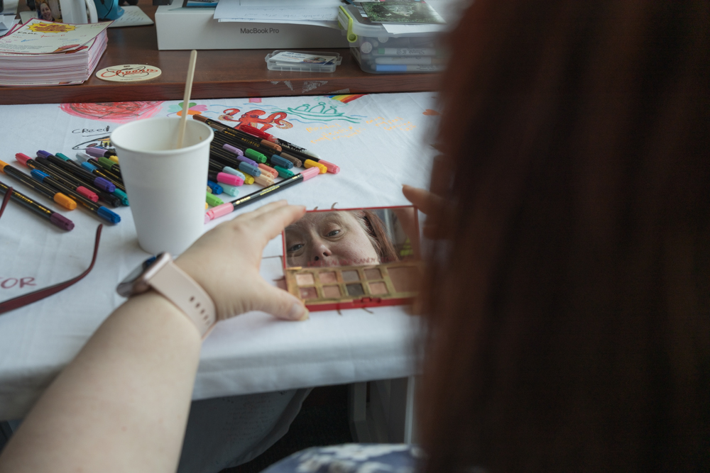 Laurie checks her makeup before her Sacred Stories interview, with a coffee cup and colorful art supplies spread across the table.